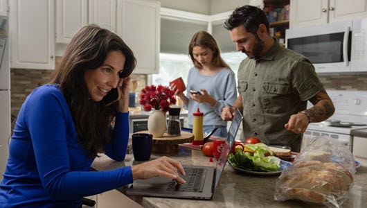 Family of three in their kitchen with mom on a laptop, daughter on her phone and dad making a sandwich.